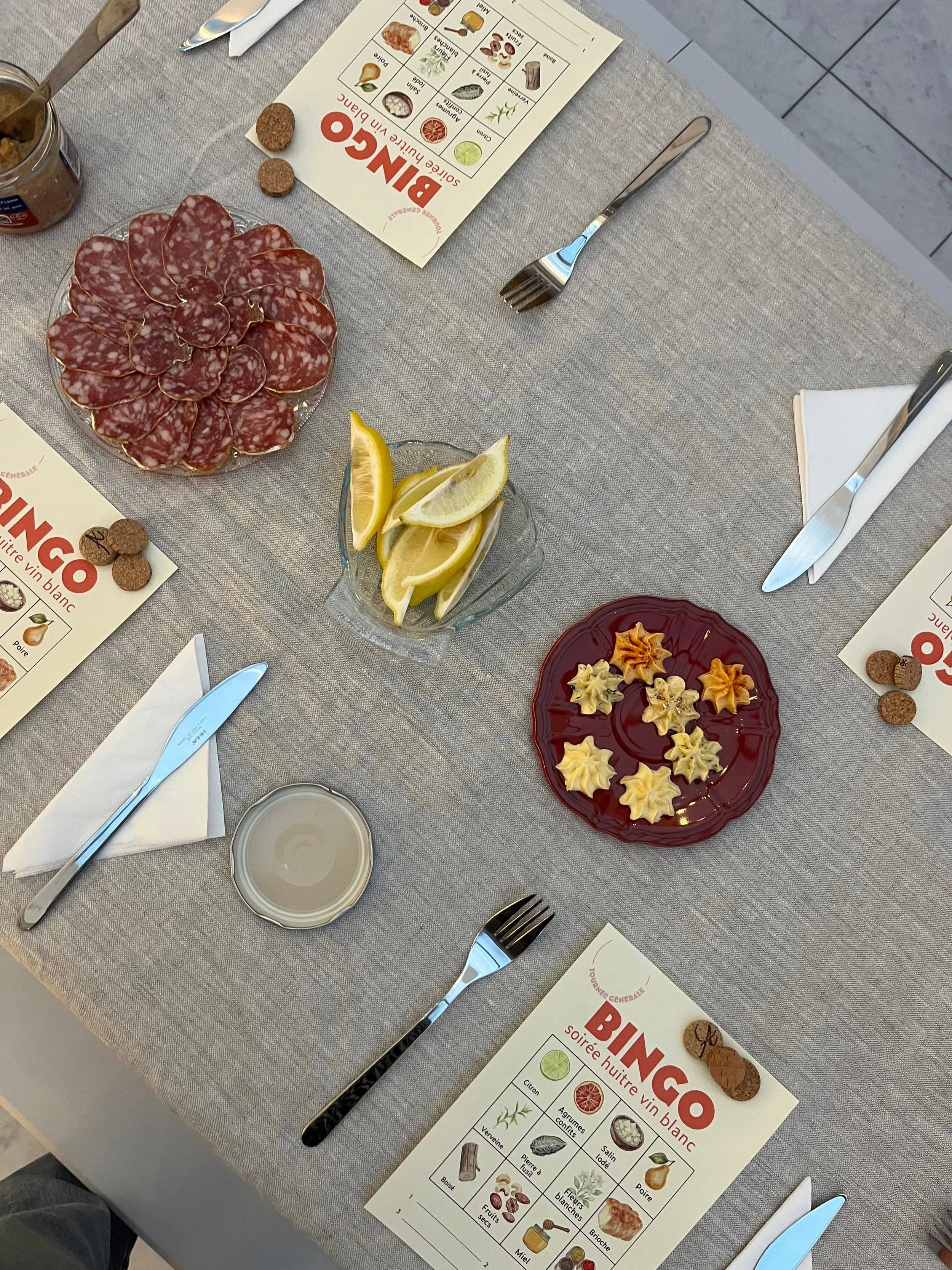 Une table de soirée à la cave vue du dessus : cartes de bingo, assiette de charcuterie, citrons et couverts