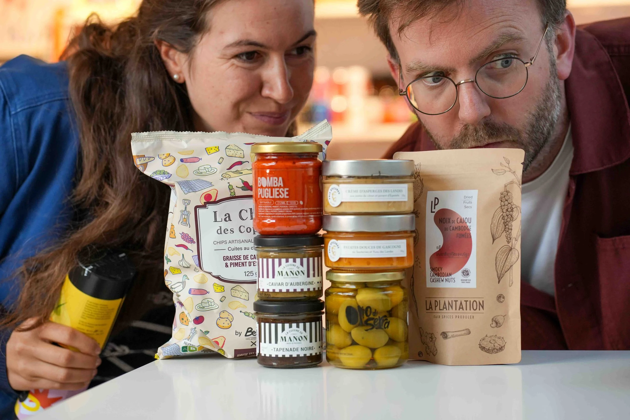 Charlotte et Alexis, l'œil gourmand, derrière une pile d'épicerie pour l'apéro : chips, tartinables, olives et noix de cajou fumées