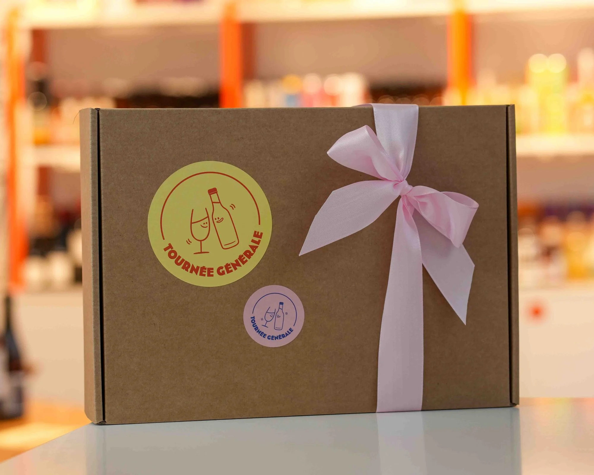 Un coffret cadeau en kraft fermé d'un ruban rose, avec les autocollants ronds Tournée Générale