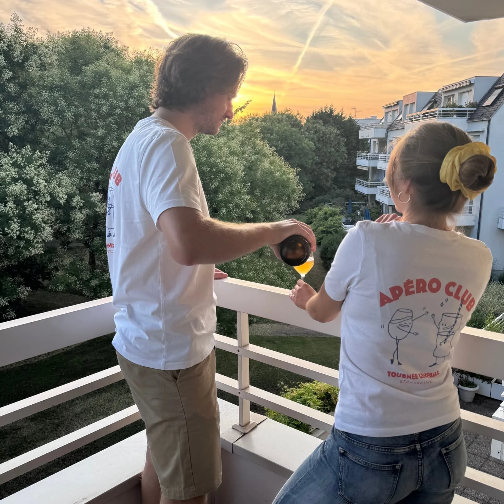 Sur un balcon au coucher du soleil, une bière blonde versée dans un verre à pied, en t-shirts Apéro Club Tournée Générale
