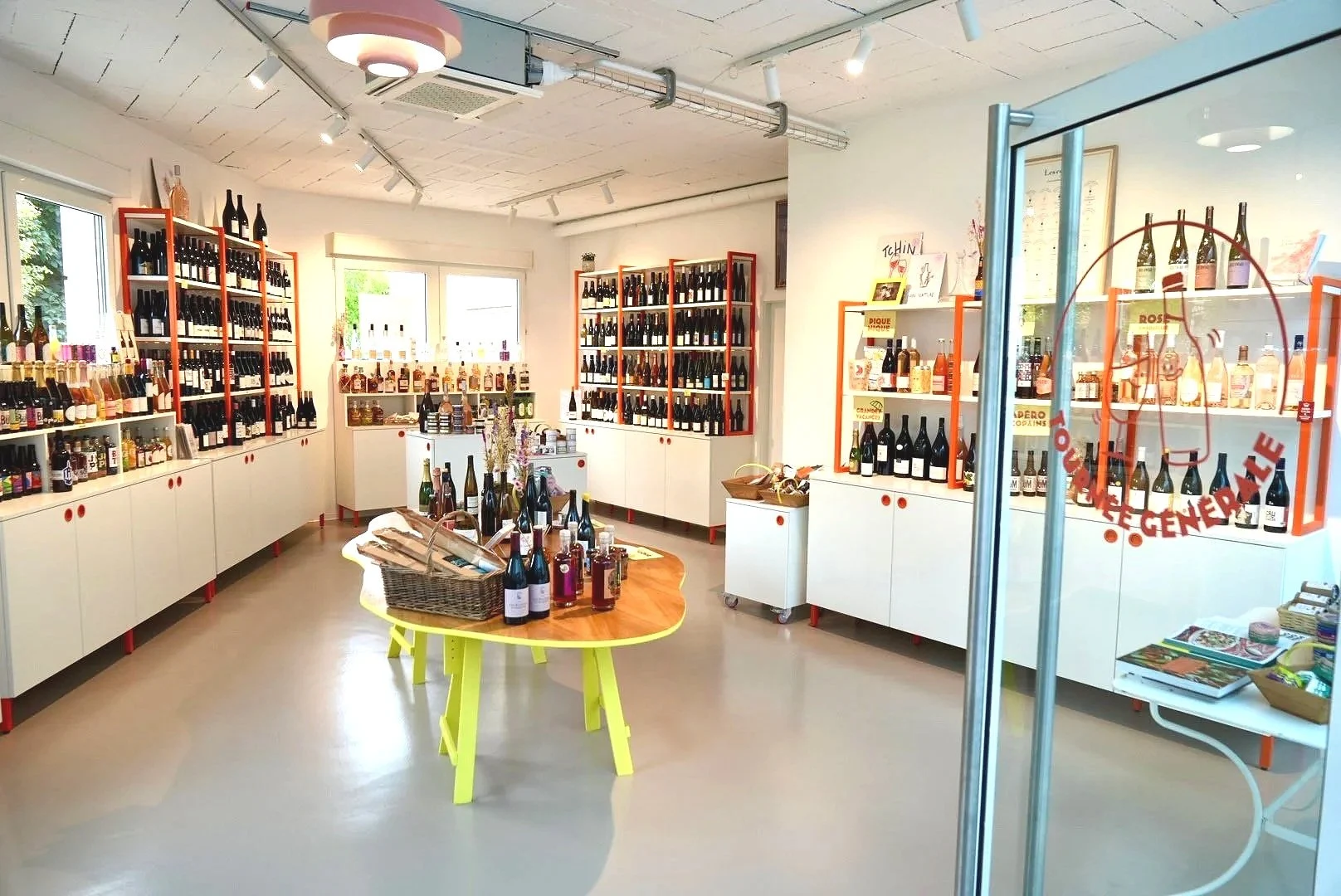 Intérieur de la cave Tournée Générale : étagères orange chargées de bouteilles et table jaune au centre