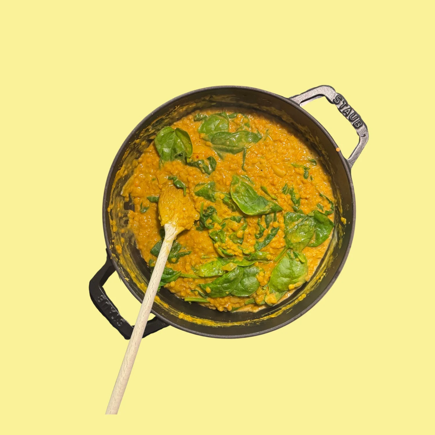 Un dahl de lentilles corail aux pousses d'épinard dans une cocotte en fonte noire, avec une cuillère en bois, sur fond jaune