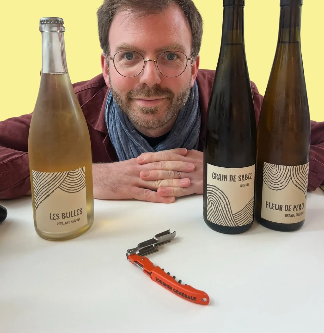 Alexis Roposte derrière trois bouteilles de vin et un tire-bouchon, sur fond jaune