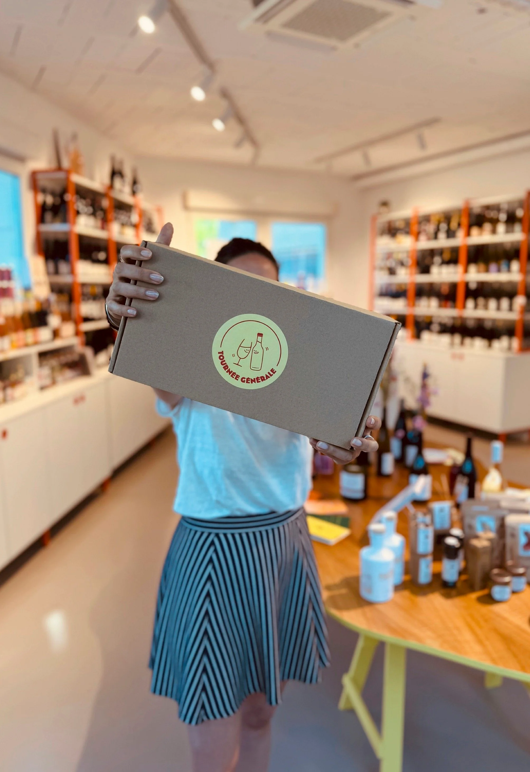 Dans la cave, une personne brandit un coffret cadeau en kraft marqué du logo rond Tournée Générale