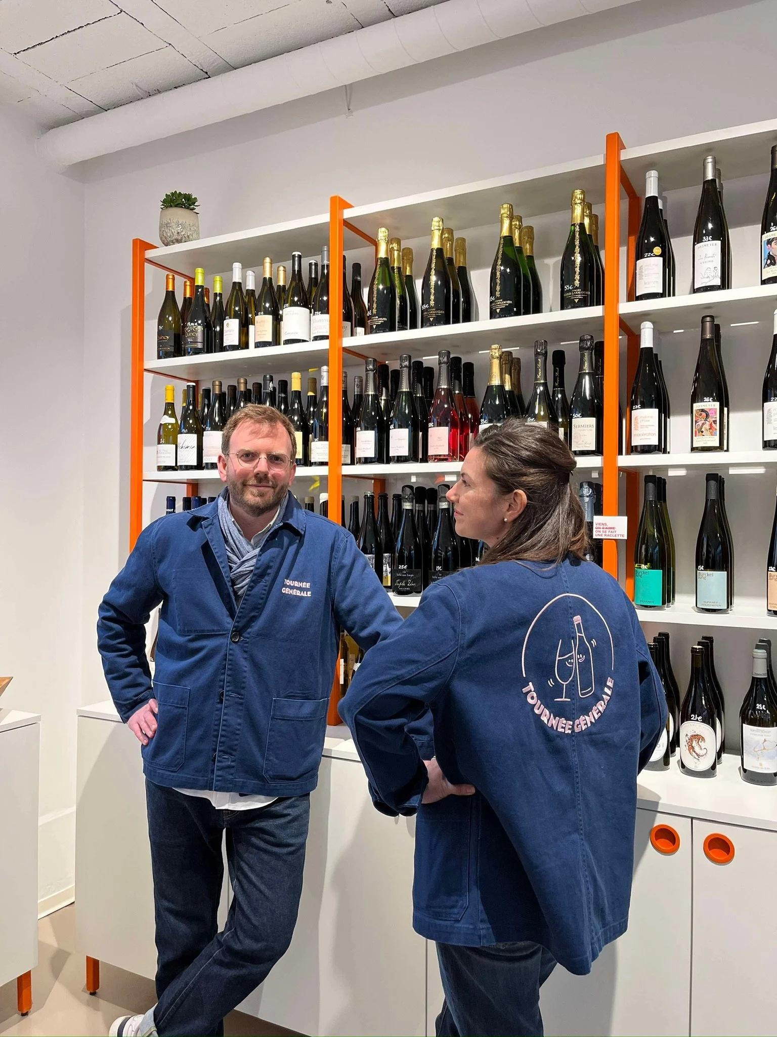 Alexis et Charlotte Roposte, frère et sœur, en vestes de travail bleu marine devant les étagères de vins de la cave