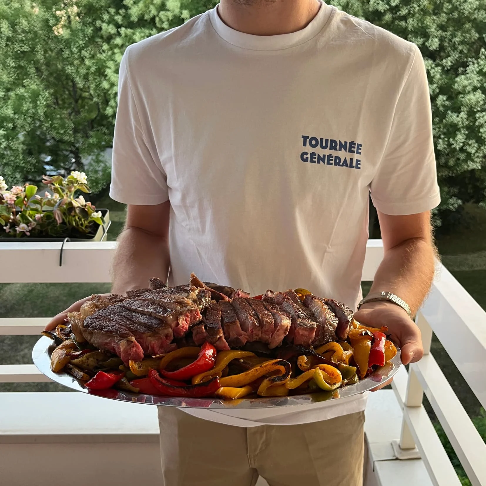 Grand plat de viande grillée tranchée sur des poivrons, tenu sur un balcon par une personne en t-shirt Tournée Générale